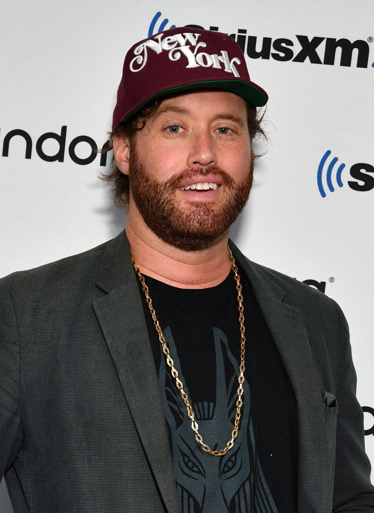 T.J. Miller is wearing a New York cap, gray blazer, and chain necklace while posing in front of a SiriusXM and Pandora backdrop