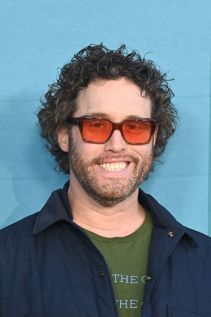 T.J. Miller smiles while wearing rectangular orange-tinted glasses, a green shirt, and a dark blue jacket