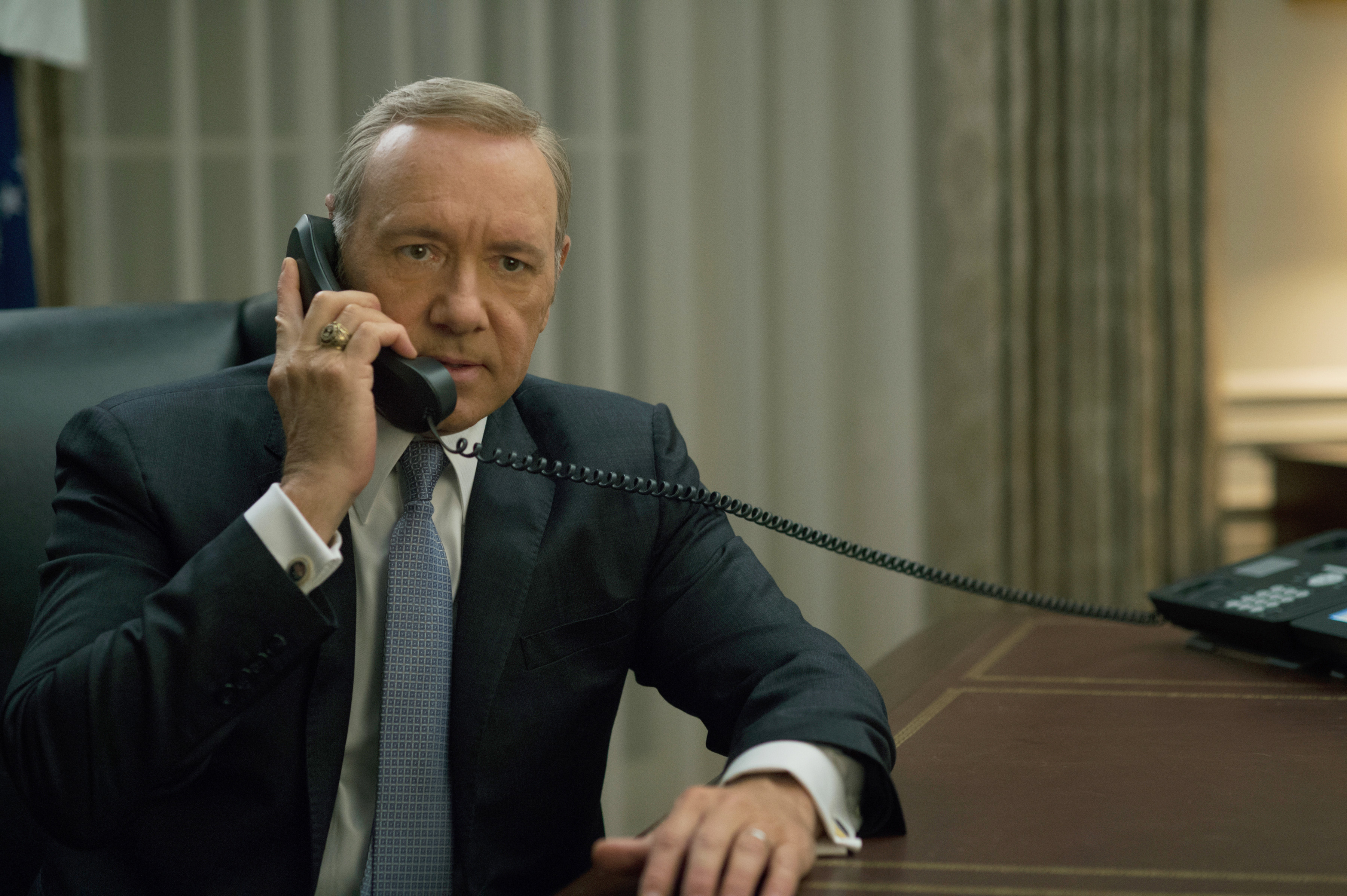 A man in a suit and tie, speaking on a corded phone while seated at a desk in an office setting