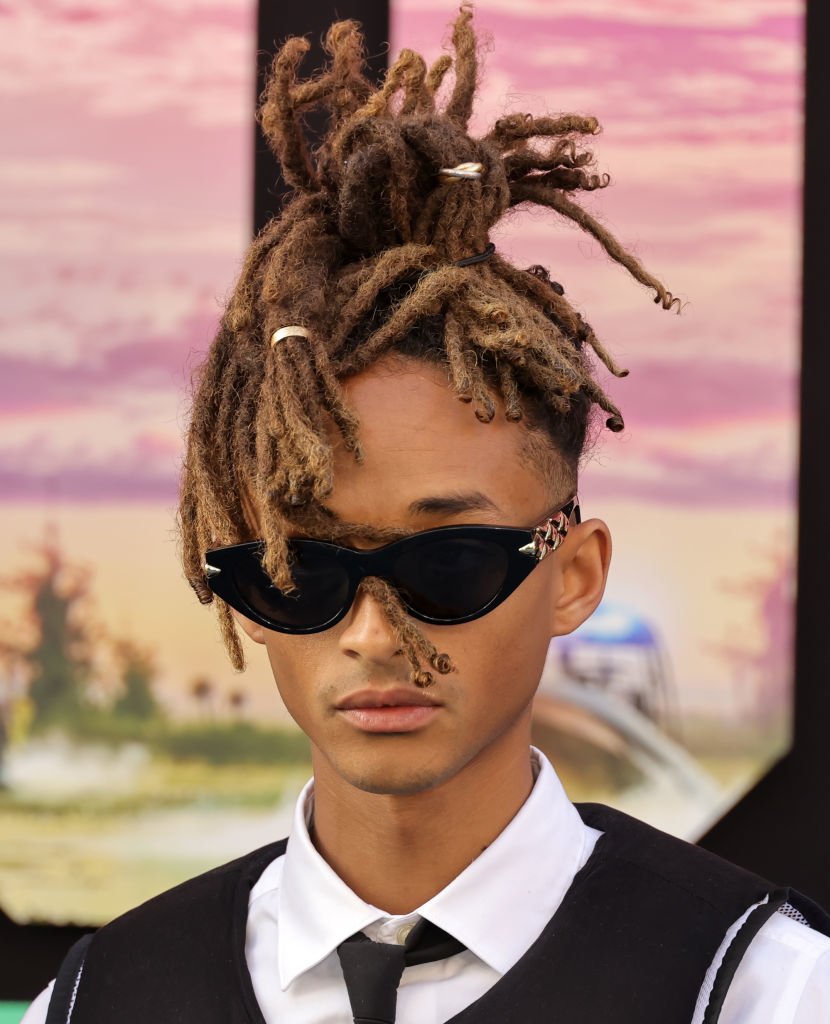 Jaden Smith wearing sunglasses and a formal black and white outfit with a tie, posing in front of a scenic backdrop