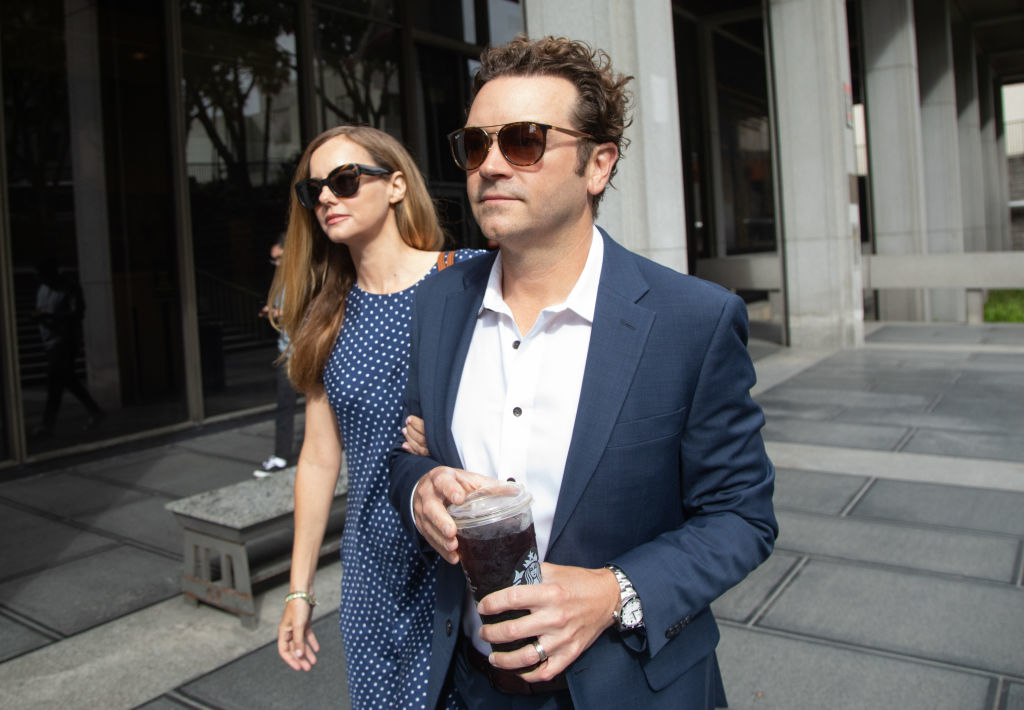 A woman in a polka dot dress and sunglasses walks arm-in-arm with a man in a suit and sunglasses holding a large drink. They appear to be outside a city building