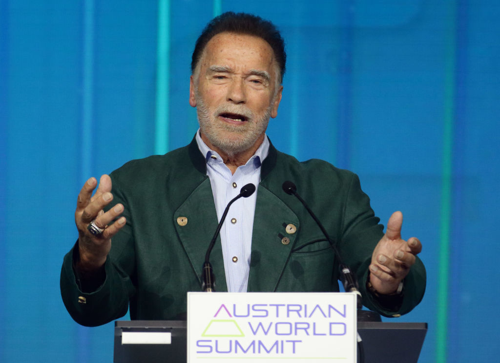 Arnold Schwarzenegger speaking at the Austrian World Summit, wearing a green jacket over a button-up shirt
