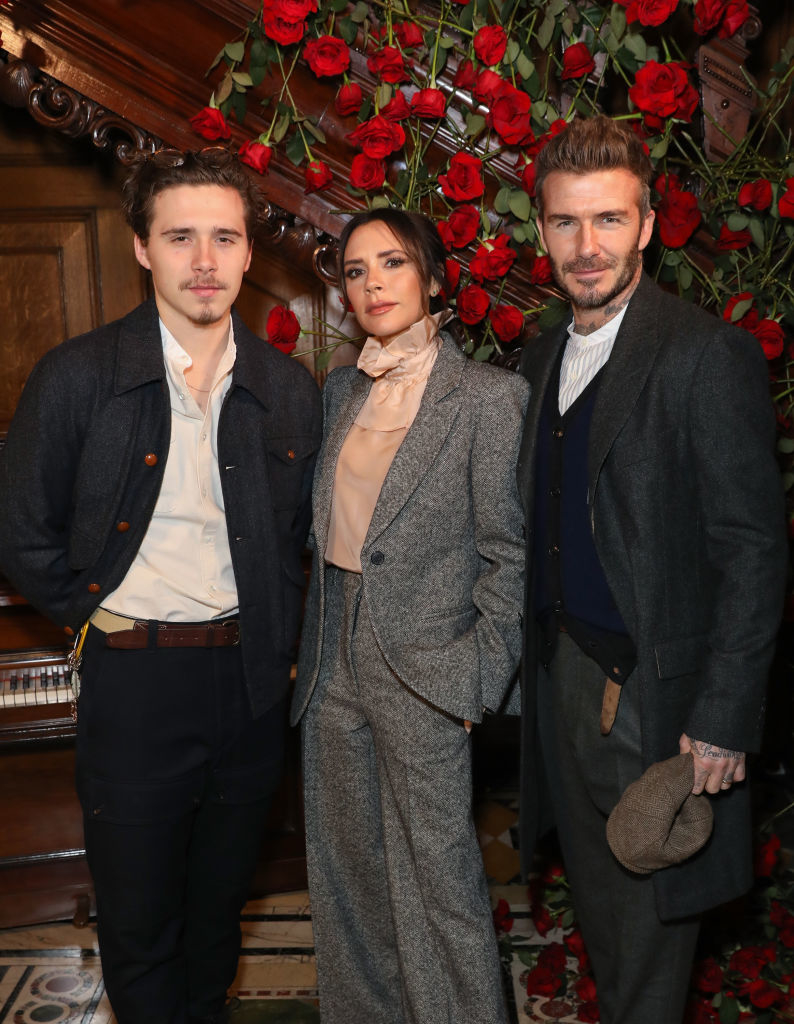 Brooklyn Beckham, Victoria Beckham, and David Beckham pose together. Brooklyn and David wear suits, and Victoria wears a stylish blazer and matching pants