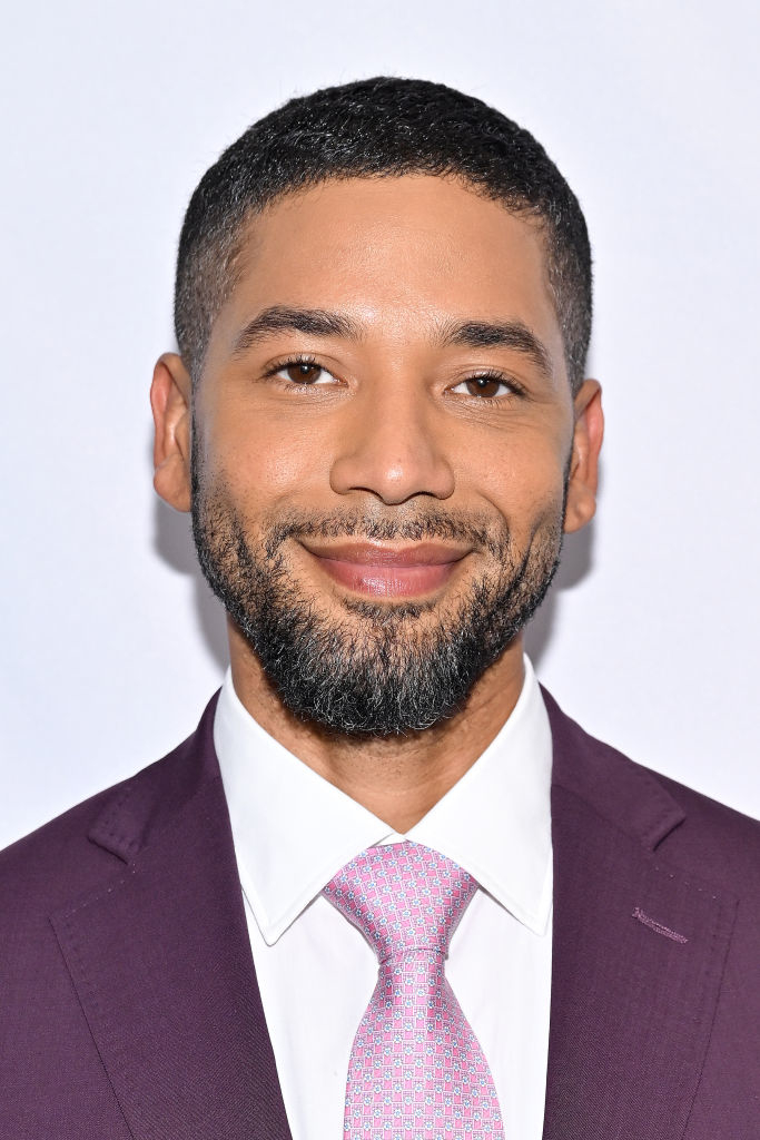 Jussie Smollett is smiling at the camera, wearing a suit with a tie