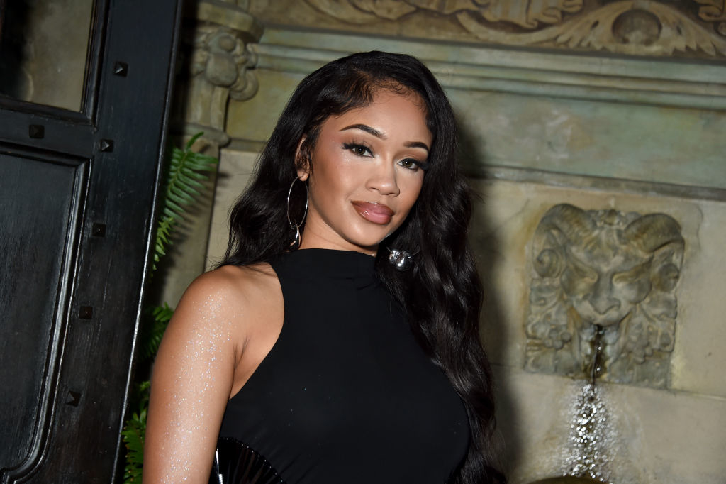 Saweetie poses in a sleeveless black dress in front of a decorative background with a lion head fountain