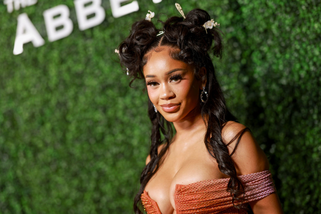 Saweetie stands in front of a greenery backdrop wearing an off-the-shoulder, structured dress with floral hair accessories