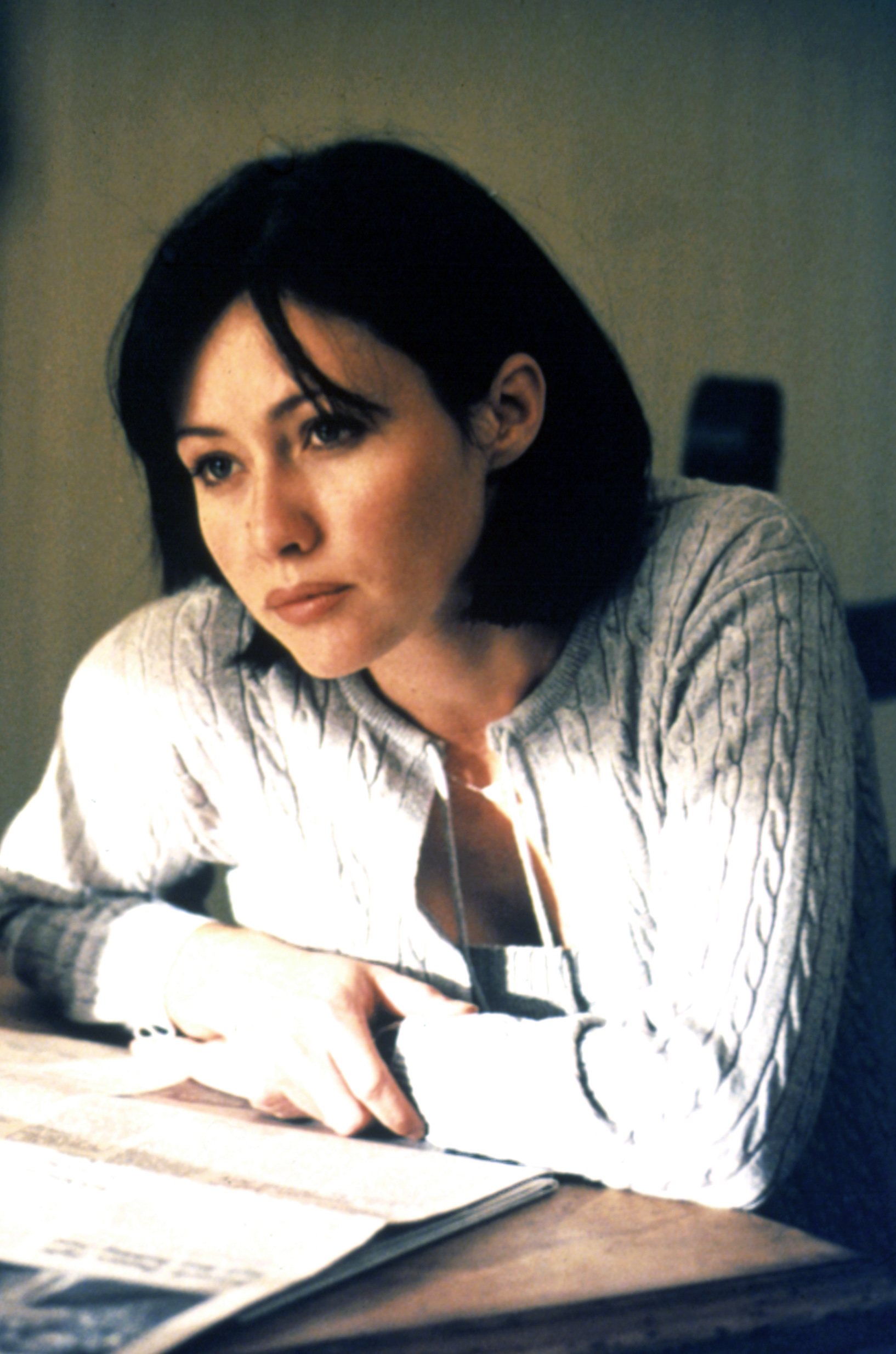 A woman with shoulder-length hair is leaning on a table, wearing a cable-knit cardigan, and looking thoughtful; her gaze is directed off-camera