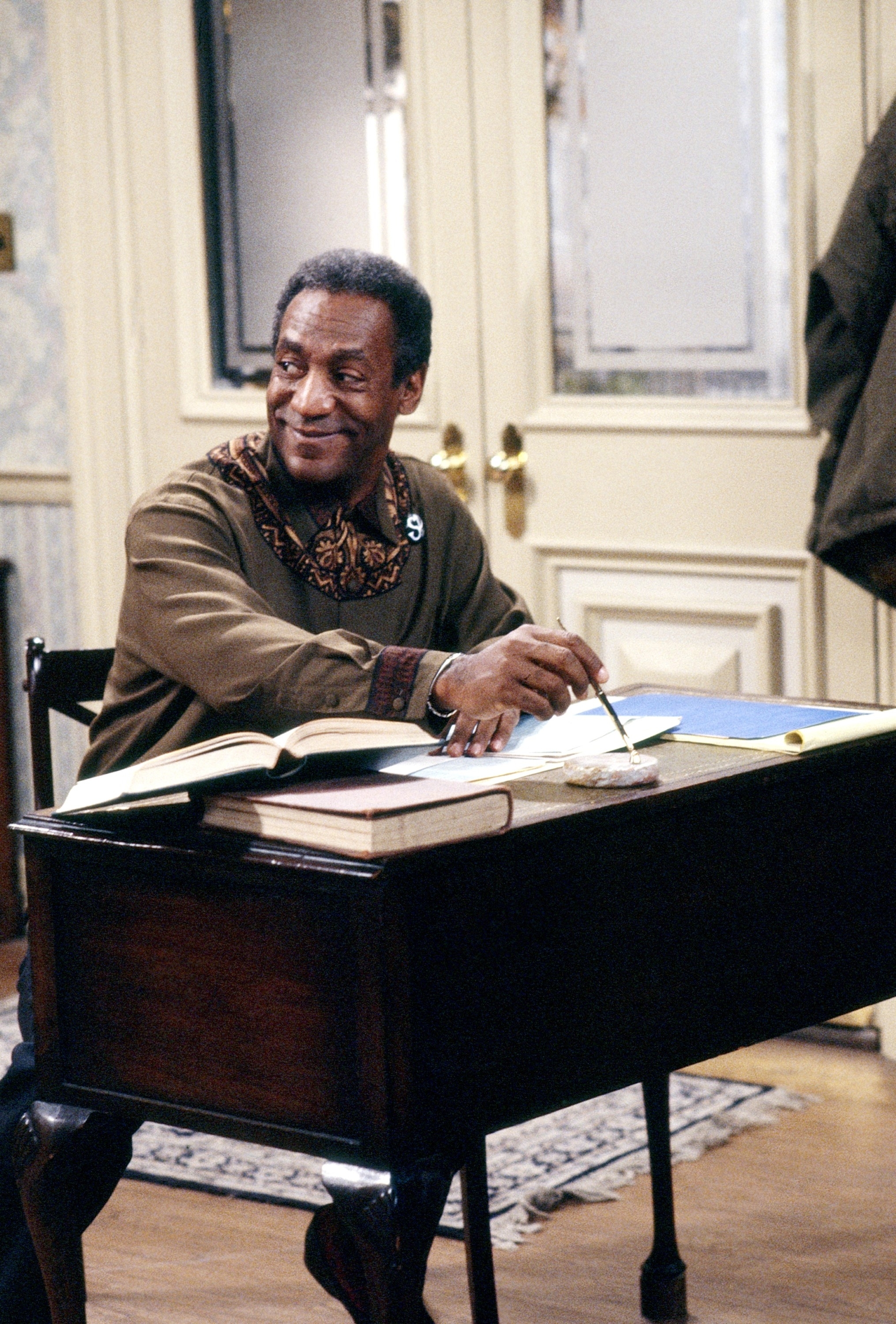 An individual sits at a desk smiling, wearing a shirt with patterned details around the collar, with an open book and a notebook on the desk