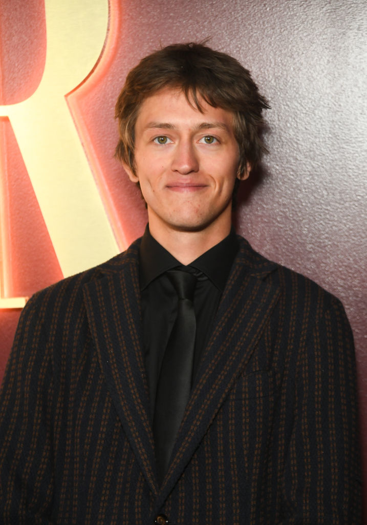 A man wearing a striped suit and black shirt stands in front of a backdrop with illuminated letters