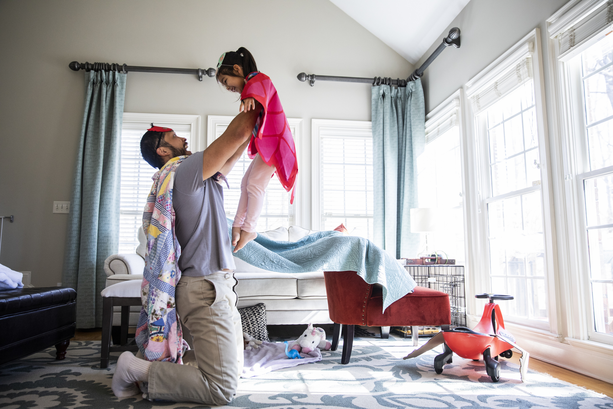 Father lifting his young daughter into the air in a bright, cozy living room with toys and blankets around