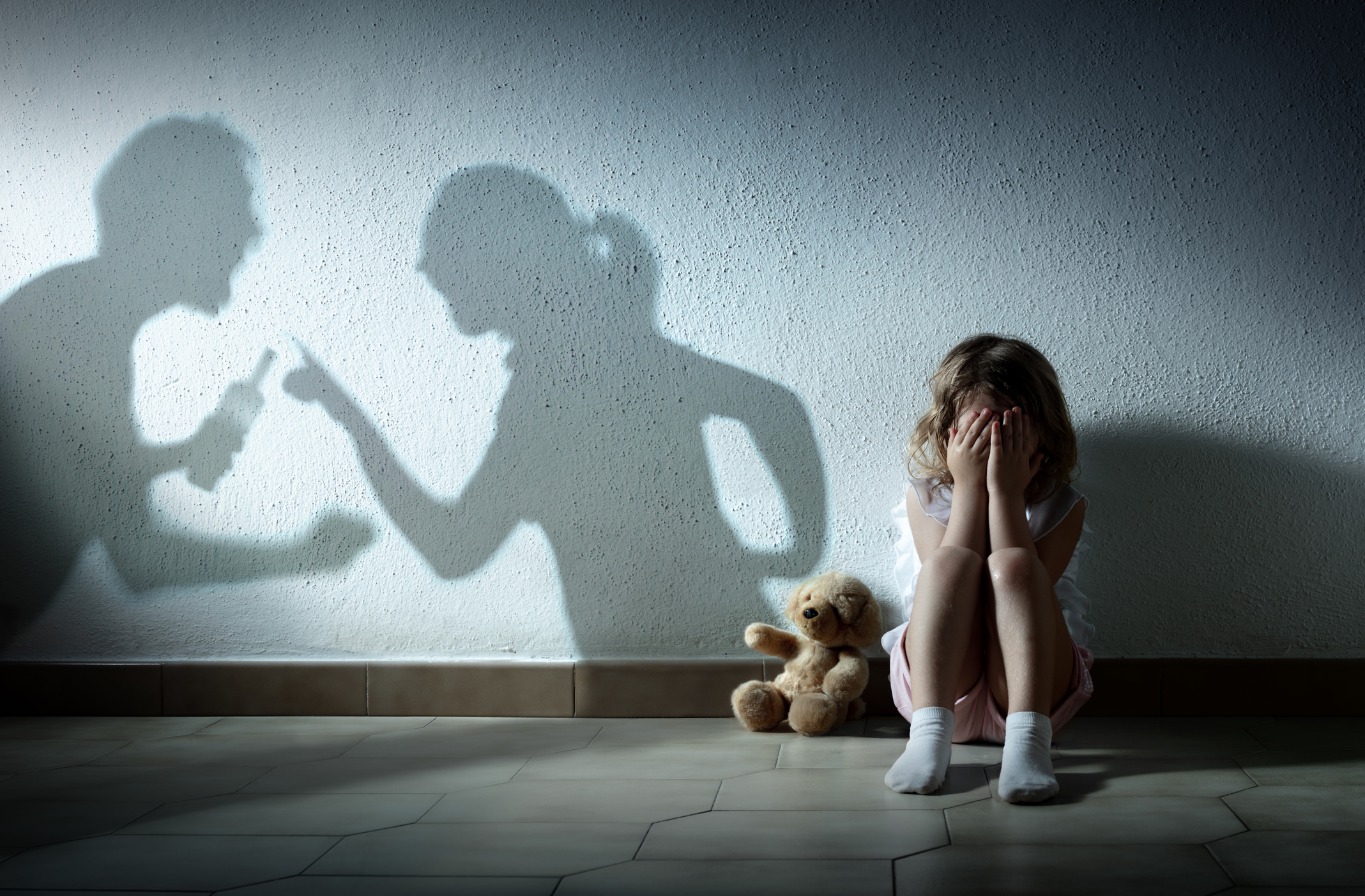 A young child sits against a wall with a teddy bear, covering their face. Shadows on the wall show two adults arguing and pointing fingers at each other