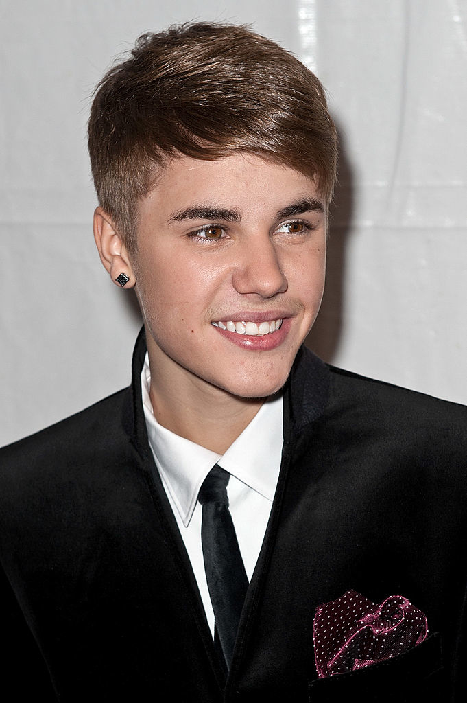 Justin Bieber smiling, wearing a black suit with a white shirt, black tie, and polka-dotted pocket square, and diamond stud earrings