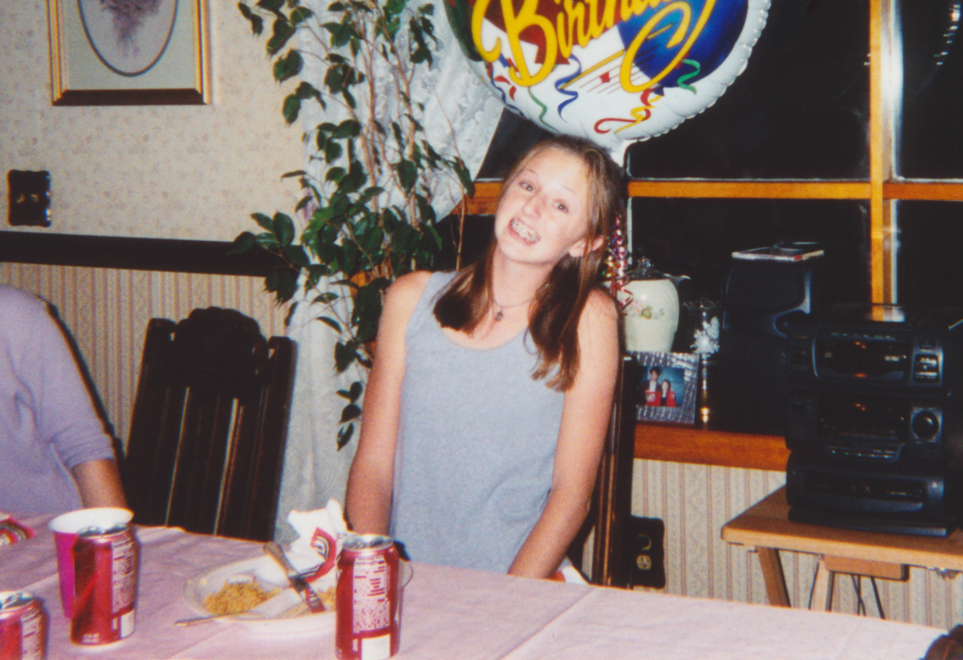 A young girl smiles at a dining table with a birthday balloon in the background. There are soda cans and a plate of food on the table