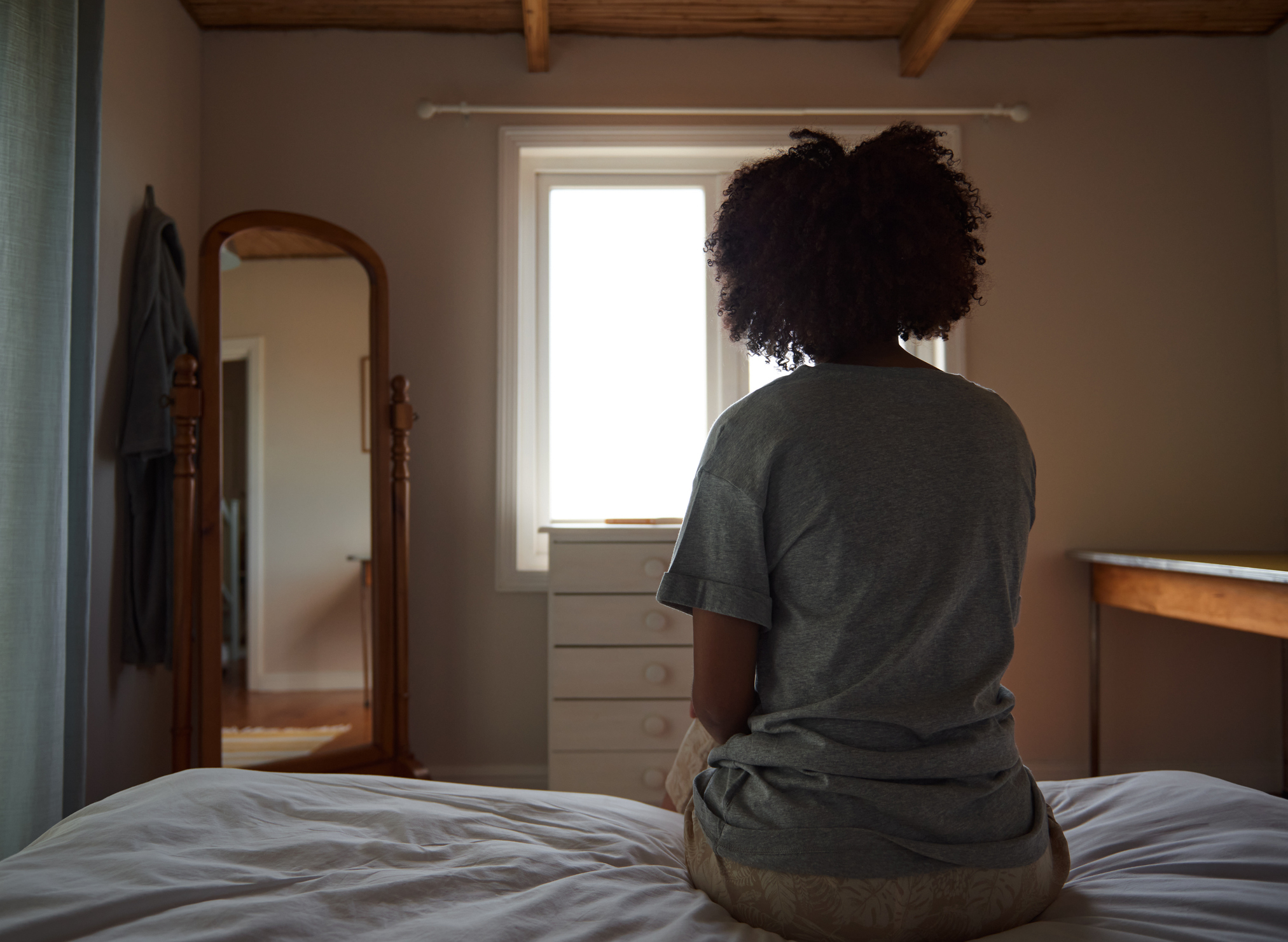 Person sitting on a bed facing a window and mirror, appearing deep in thought in a cozy bedroom