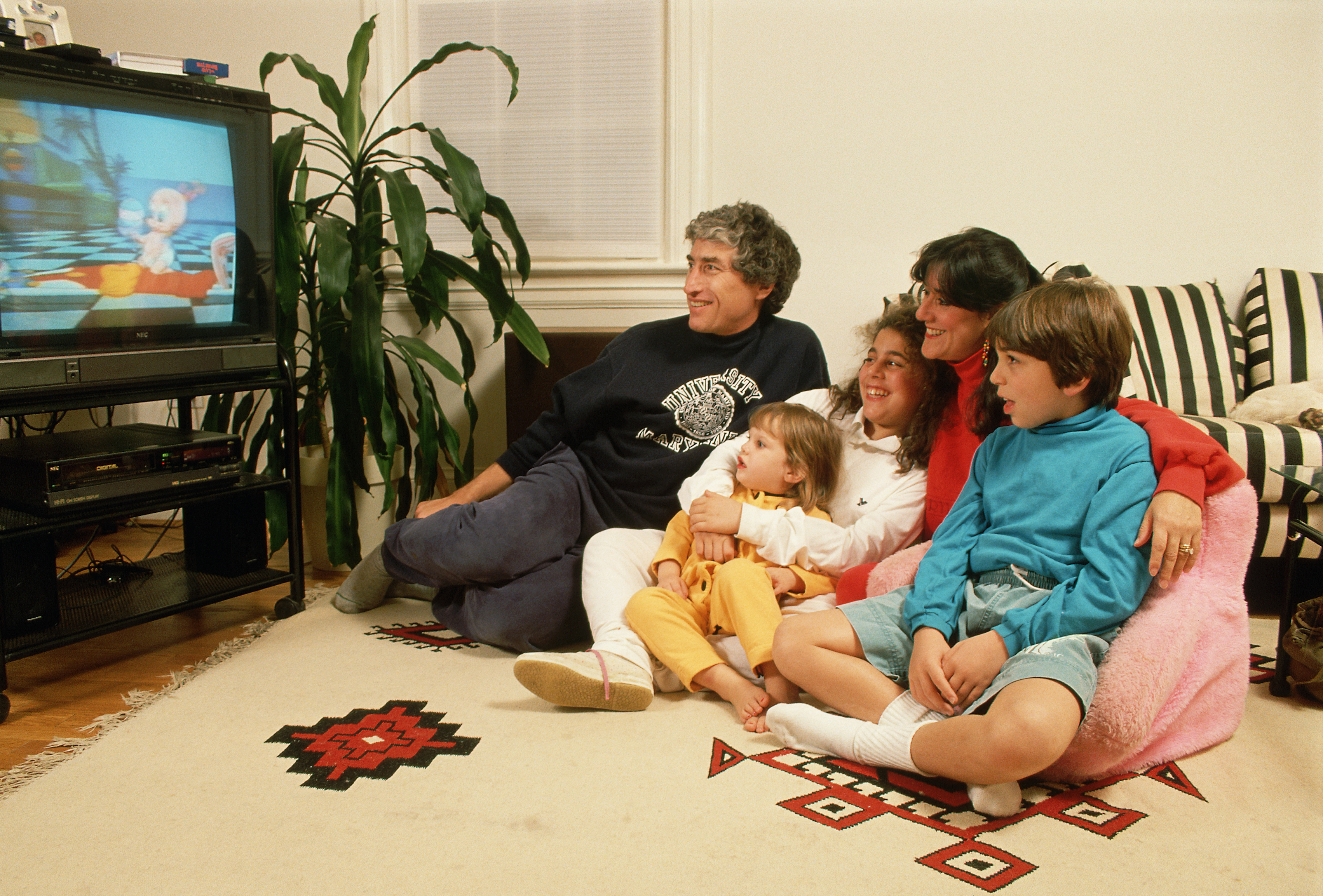 A family of five sits on a living room floor watching television together, smiling and enjoying a TV show