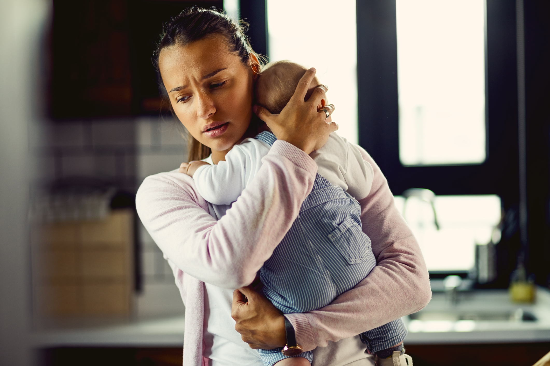 A woman holds a baby close to her chest while looking worried and concerned, standing in a kitchen