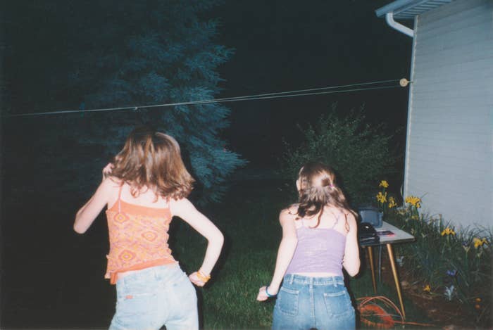 Two unidentified women are dancing outside at night next to a house with yellow flowers and a table with items on it. Both are wearing casual tops and jeans