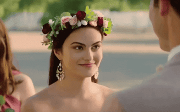 Camila Mendes, wearing a flower crown and elegant earrings, smiles and looks at a person in front of her. The background shows blurred greenery and another person