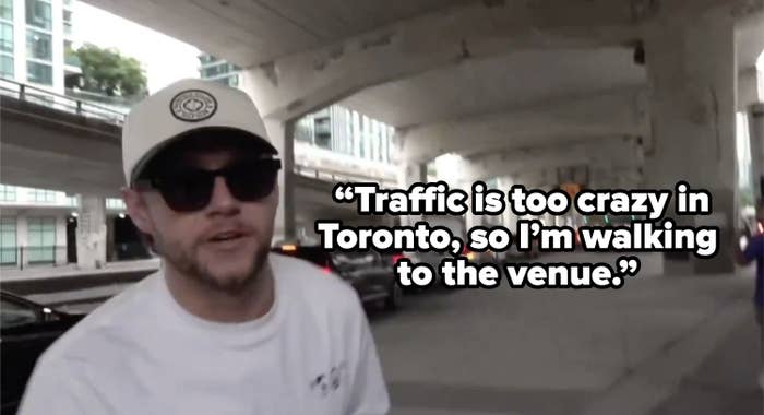 Niall Horan wears a cap and sunglasses, walking under an overpass with a line of cars in the background