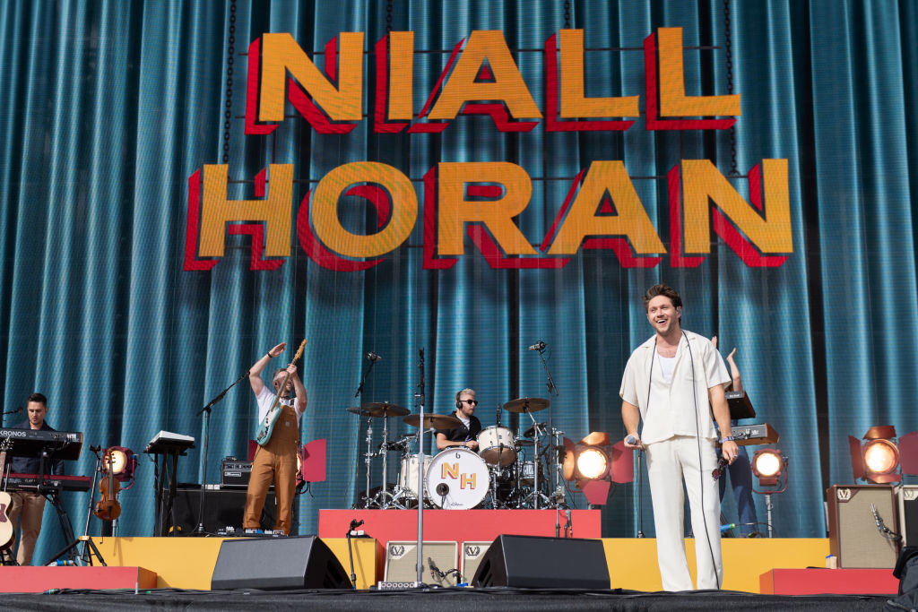 Niall Horan performs on stage with his band, featuring a backdrop of his name in bold letters. He is dressed in a casual, light-colored outfit