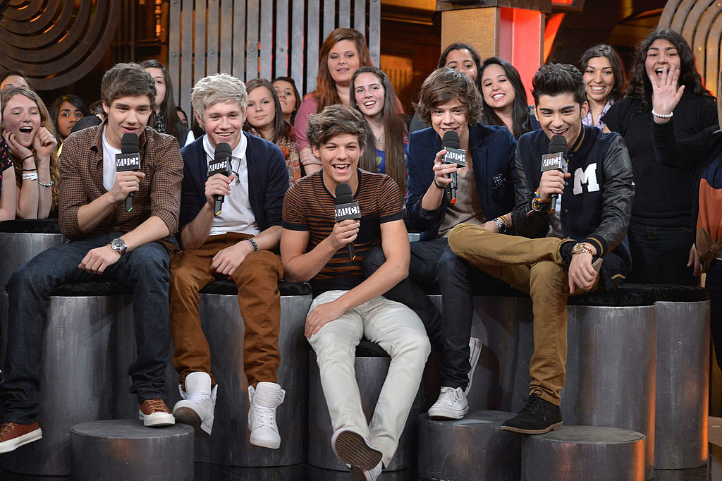 One Direction (Liam Payne, Niall Horan, Louis Tomlinson, Harry Styles, Zayn Malik) smiling and holding microphones, seated with fans in the background