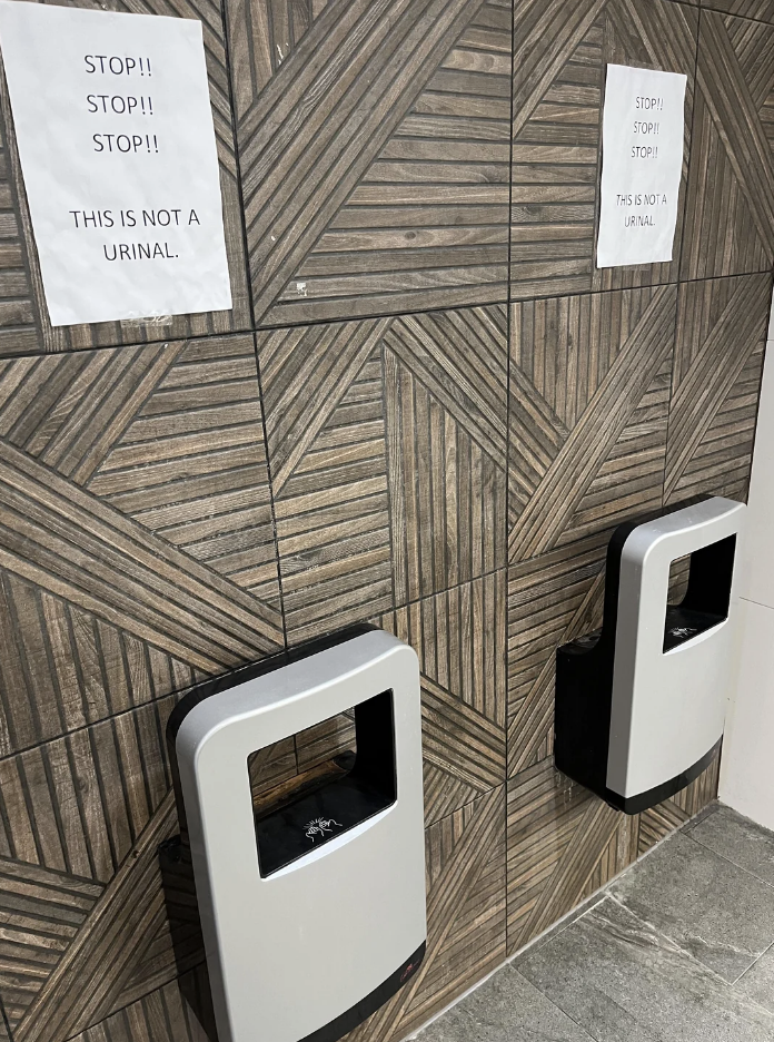 Two hand dryers on a tiled wall with signs above them stating, &quot;STOP!! STOP!! STOP!! THIS IS NOT A URINAL.&quot;