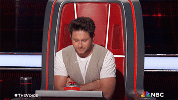Niall Horan is sitting in a red chair and pressing a button on &quot;The Voice&quot; set, with the hashtag #TheVoice and the NBC logo visible in the bottom corners