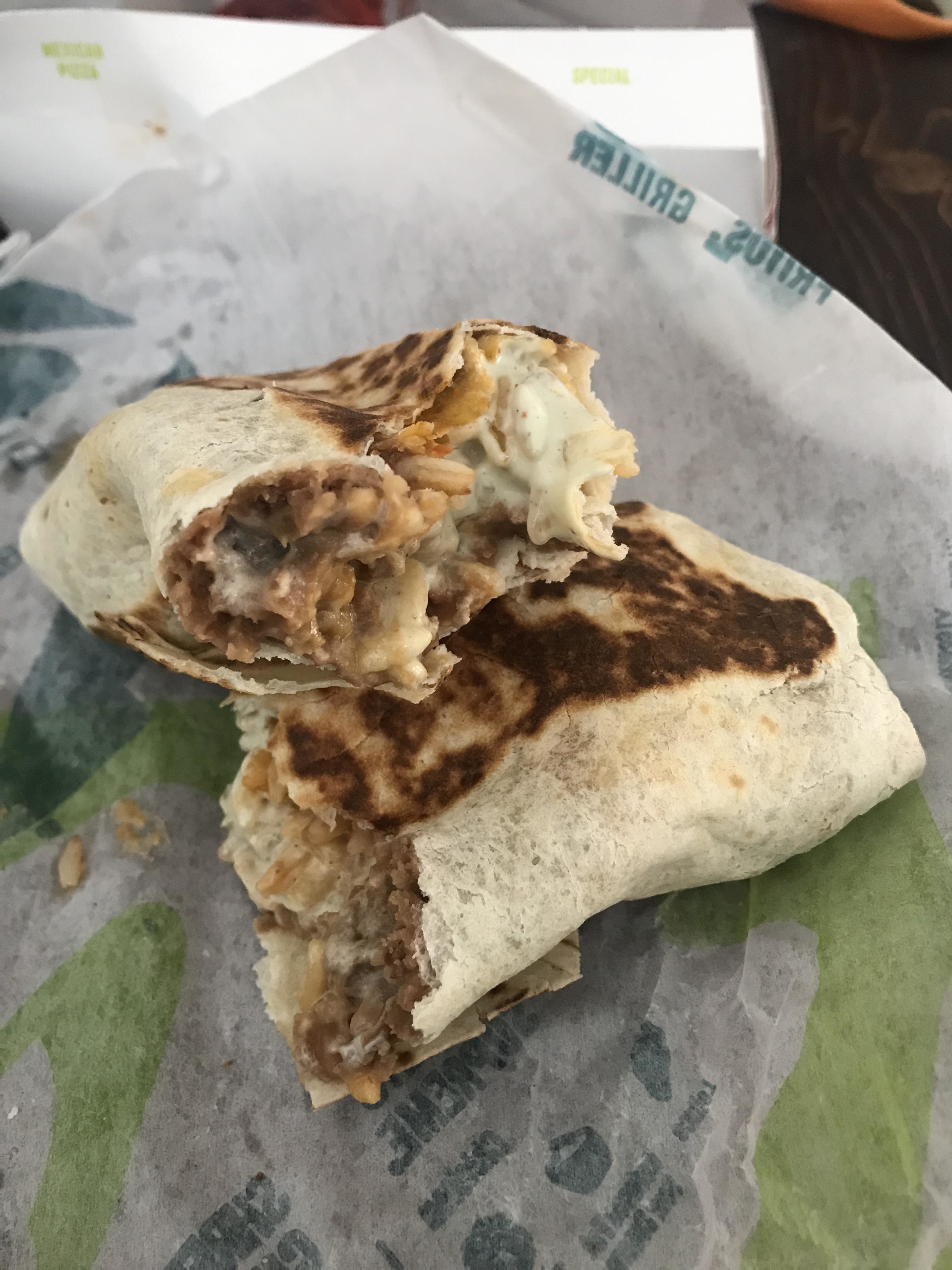 A close-up image of a half-eaten grilled burrito, showing its meaty and cheesy filling. The burrito is placed on a wrapper with text on it
