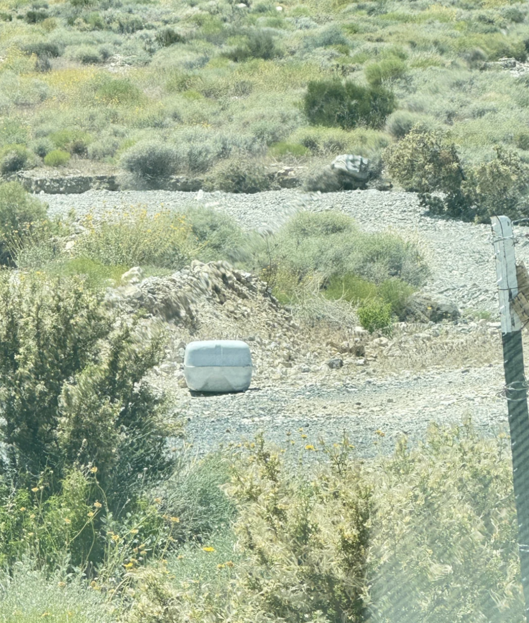 A large cube-shaped concrete block in a dry, rocky landscape with sparse vegetation and bushes in the background
