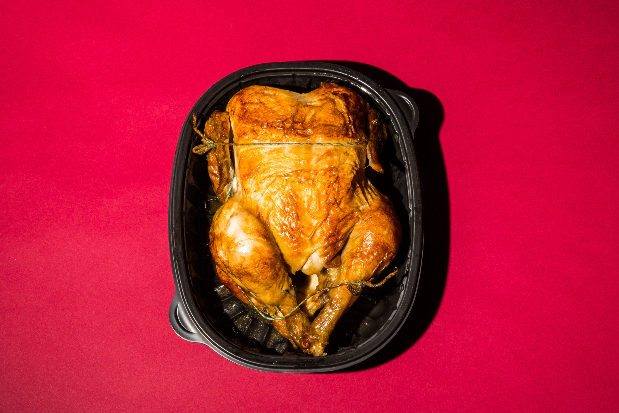 A rotisserie chicken in a black plastic container, set against a plain background