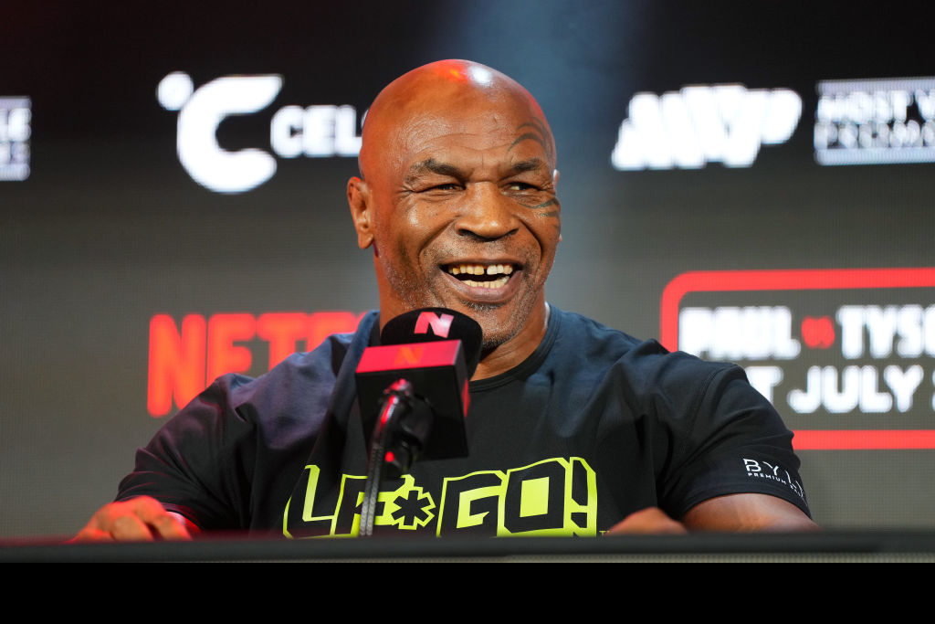 Mike Tyson at a press event, wearing a black shirt with &quot;Lift the Game!&quot; print and smiling while speaking into a microphone