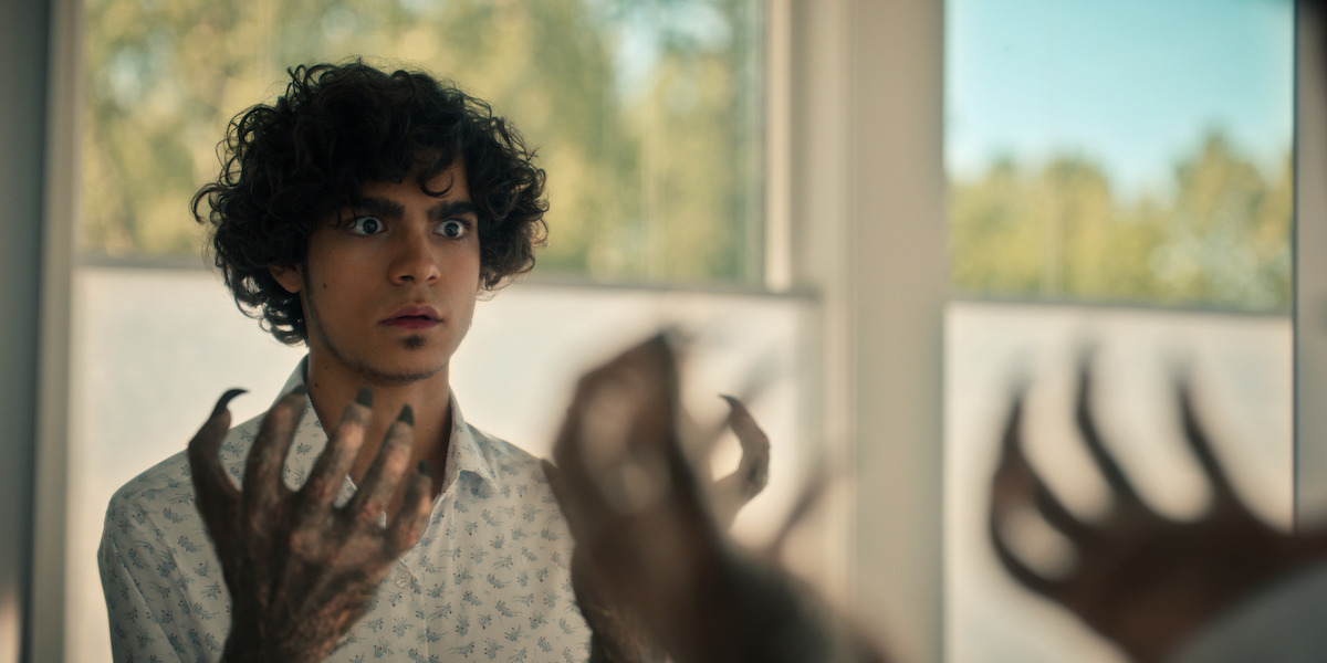 Young man with curly hair and a shocked expression looks at his hands, which have sharp claws and fur. He appears to be in front of a window with a nature scene outside