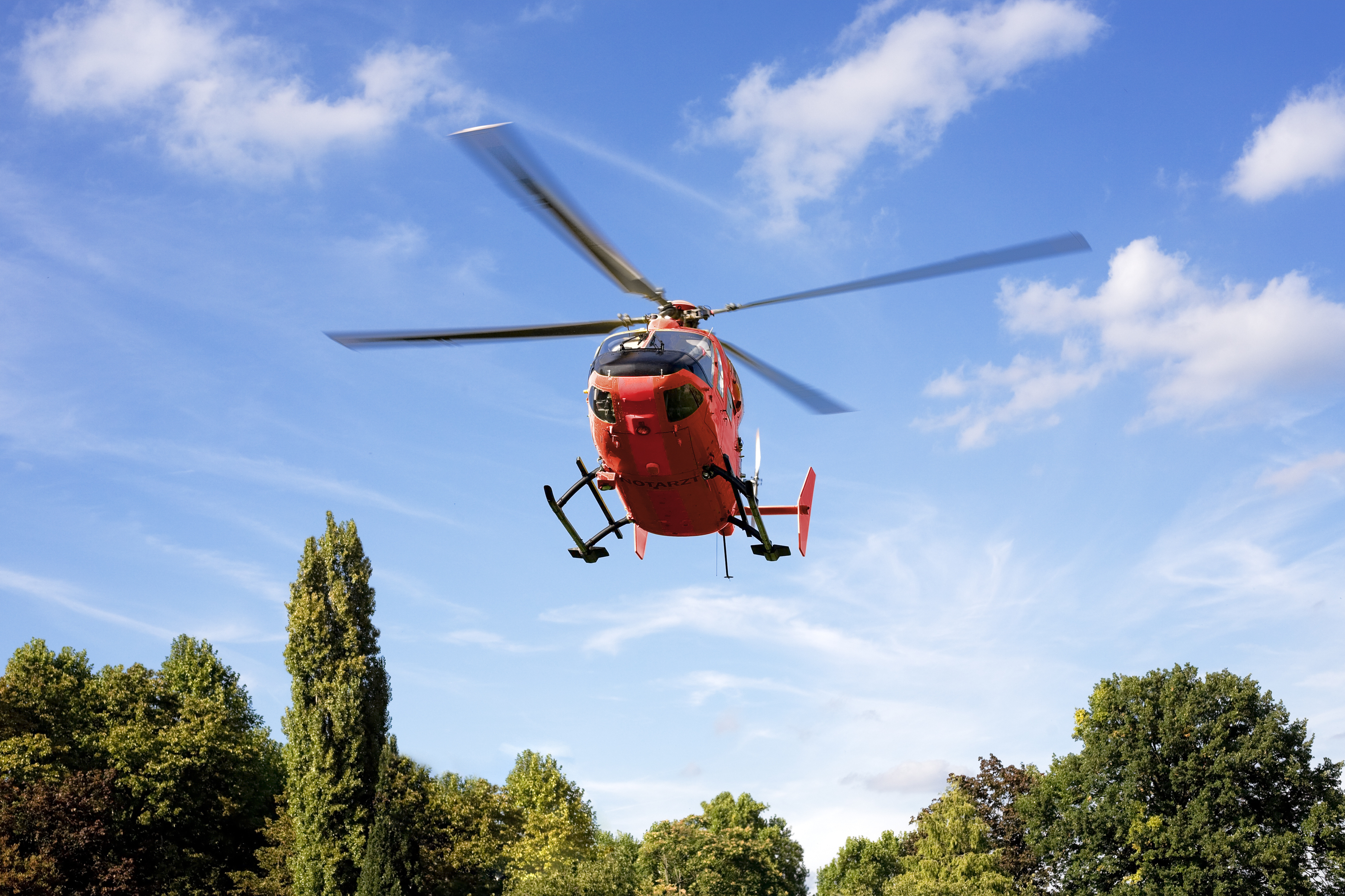 Helicopter flying over a tree-lined area against a blue sky with scattered clouds