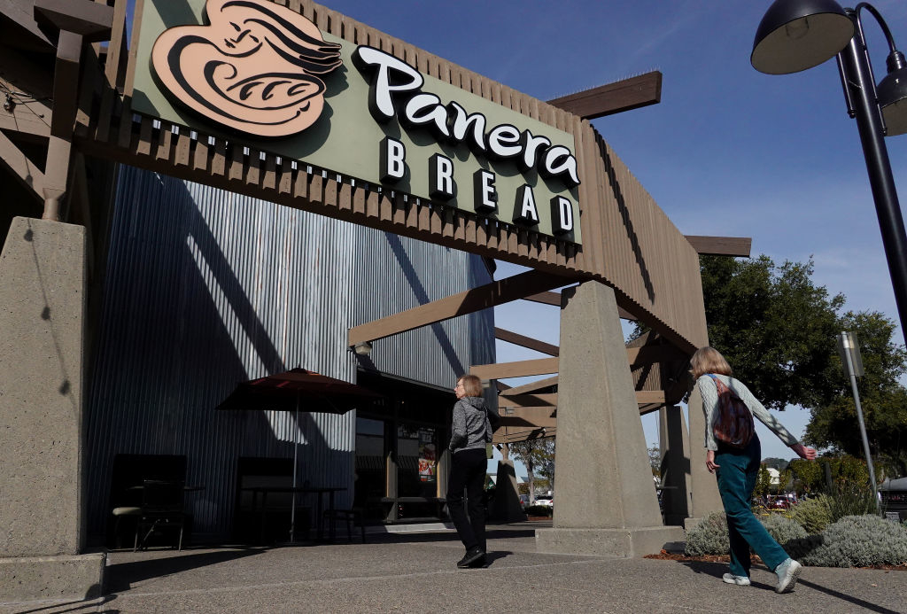 Two people walk towards a Panera Bread restaurant entrance, which has a large sign above. The photo seems to capture a casual entry scene
