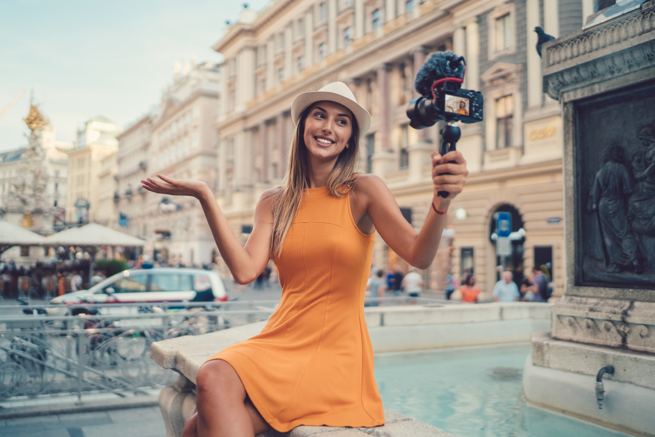 A woman in a sleeveless dress and hat smiles while holding a camera on a handheld tripod. She is seated on the edge of a fountain in an urban setting