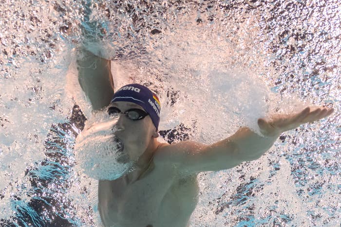 A swimmer with a dark swim cap and goggles, mid-stroke underwater, creating splashes and bubbles around the face