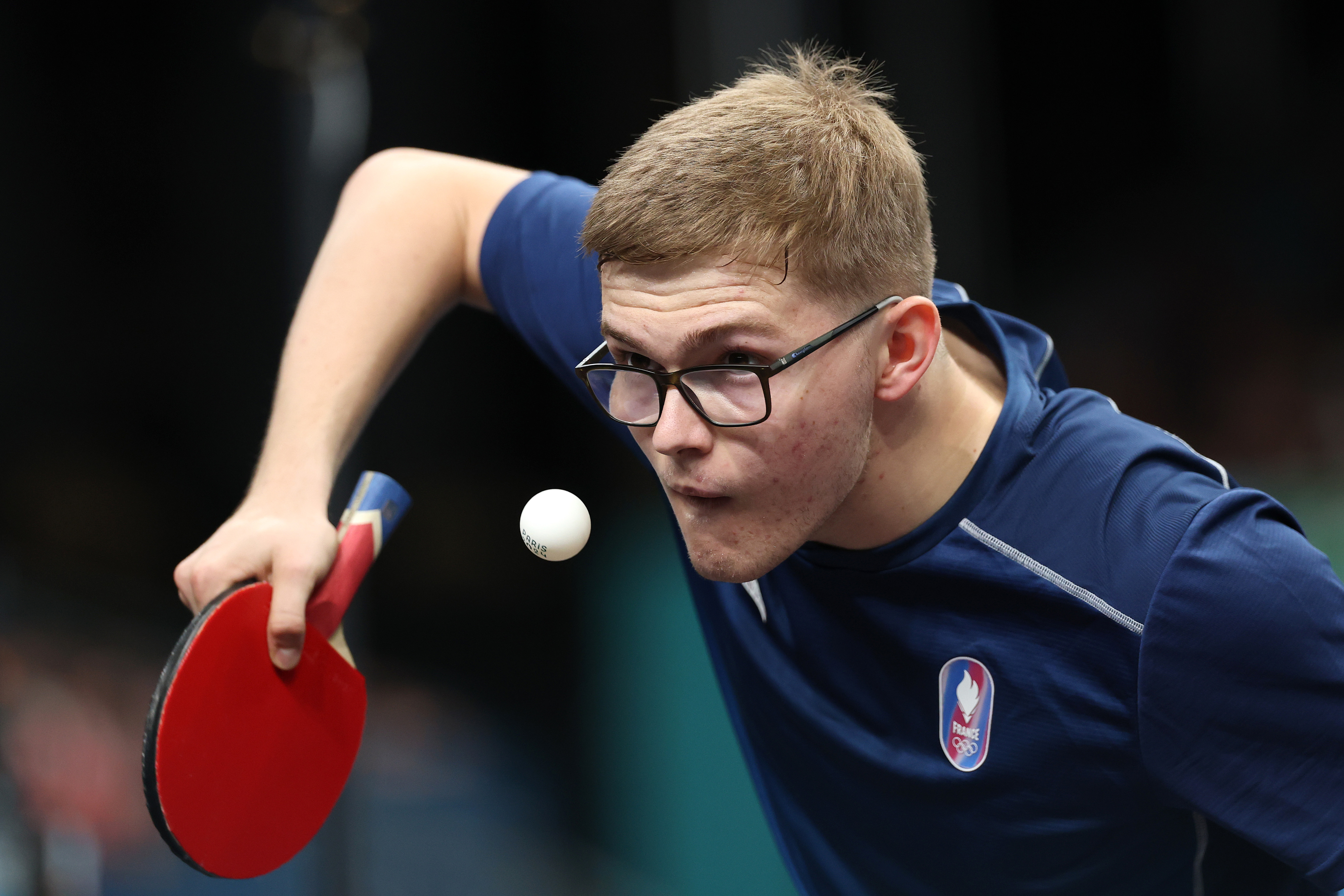 A young man with short hair and glasses intensely focuses while playing table tennis, hitting a ball with a red paddle