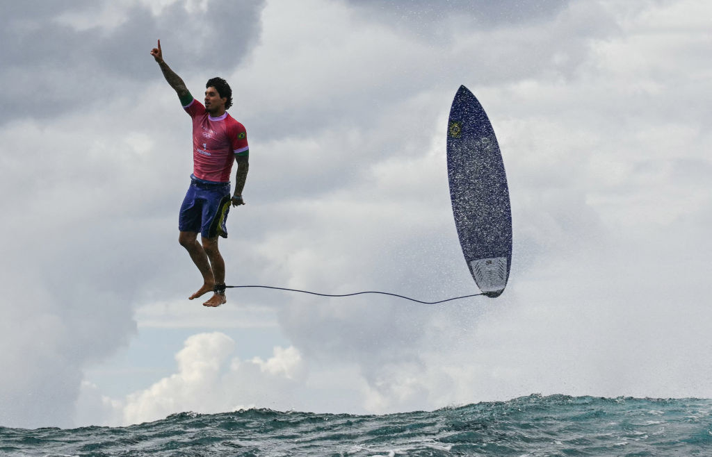 A surfer mid-air next to his surfboard above a wave, giving a raised thumb gesture with his right hand