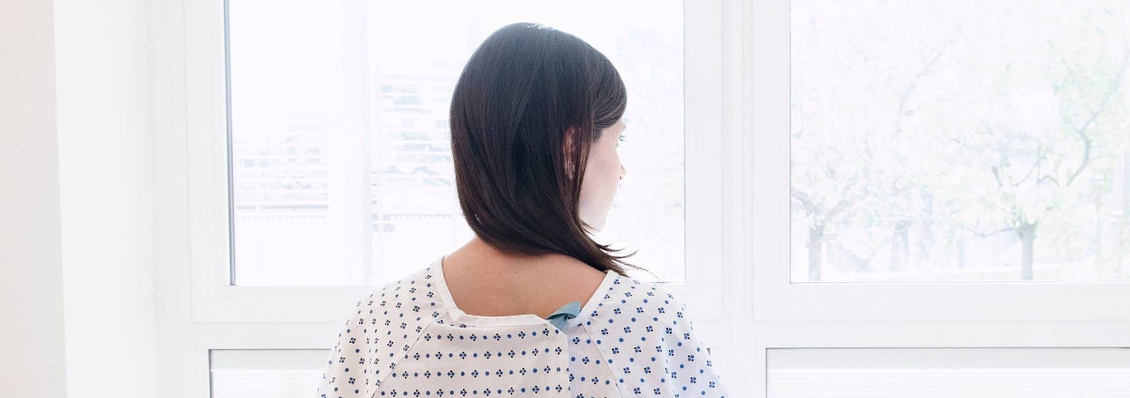 A person in a hospital gown sits on a bed, looking out the window, their back to the camera, suggesting a moment of contemplation or recovery