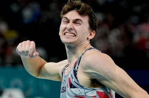 An Olympic gymnast clenches his fist and makes a determined face during a competition, wearing a gray and red uniform with USA on it