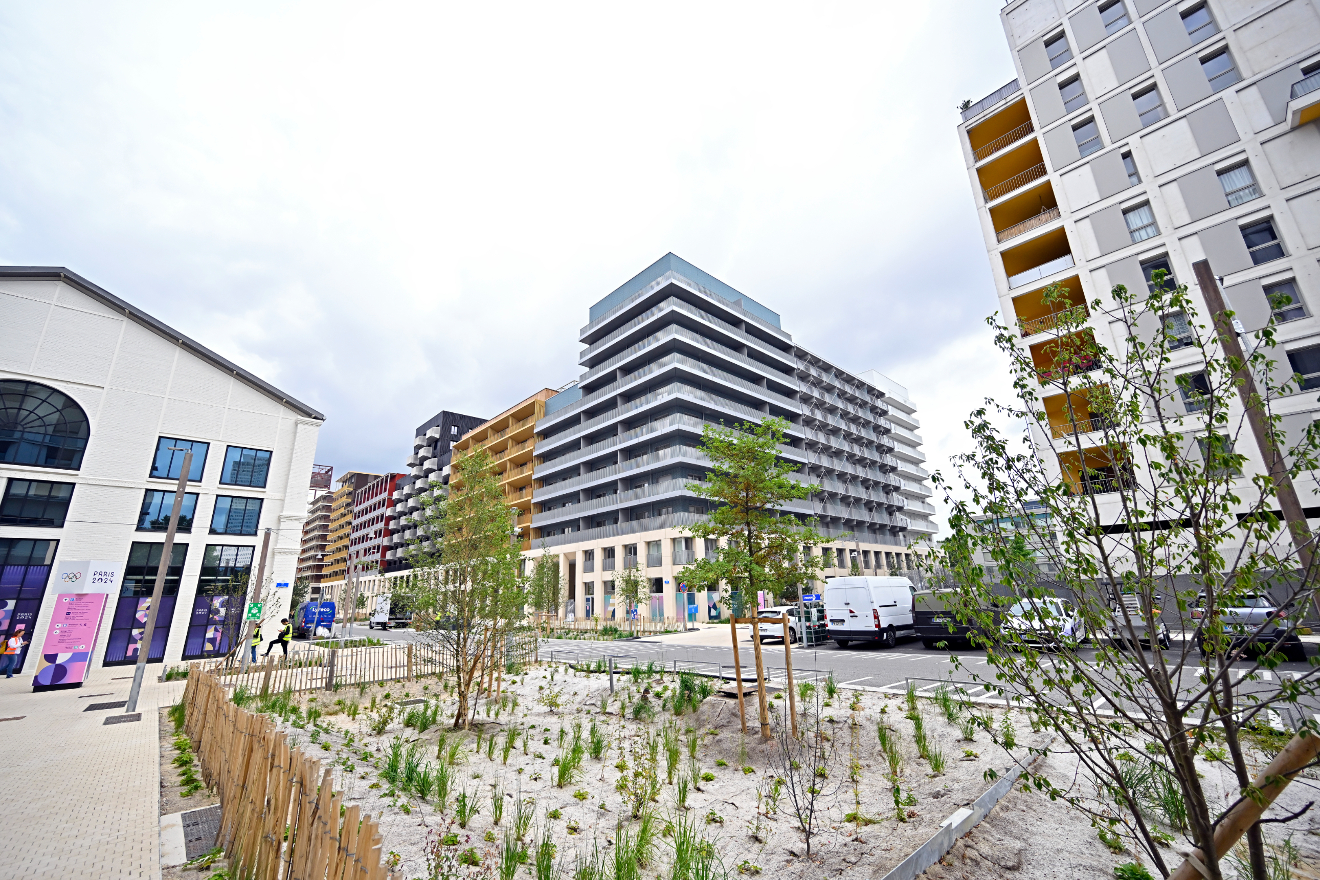 Outdoor scene showing a section of the 2024 Olympic Village featuring modern apartment buildings, small landscaped area with plants and trees, and parked vehicles on the street