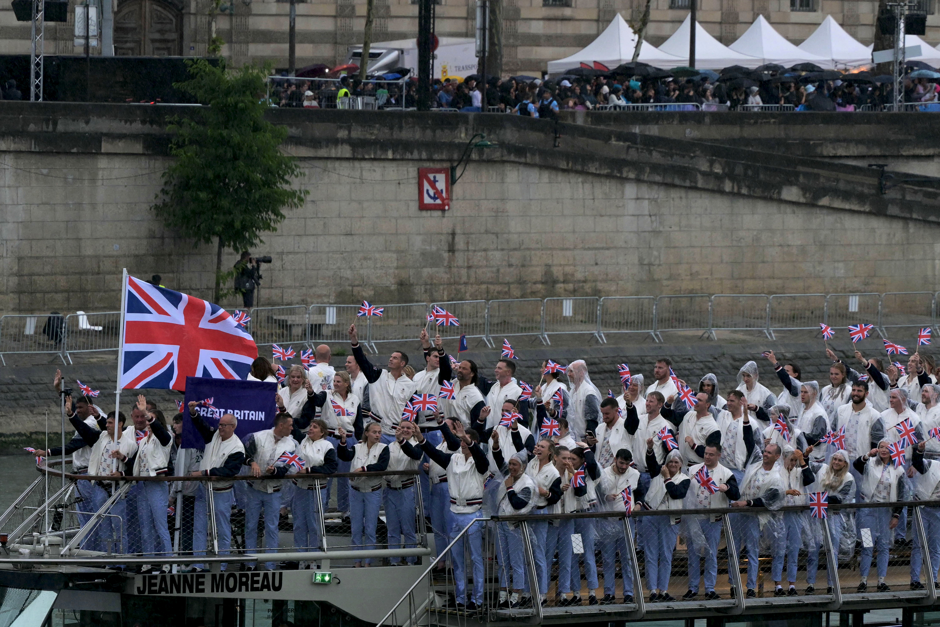 British athletes on a boat wave Union Jack flags during an outdoor event, with spectators in the background along a riverside