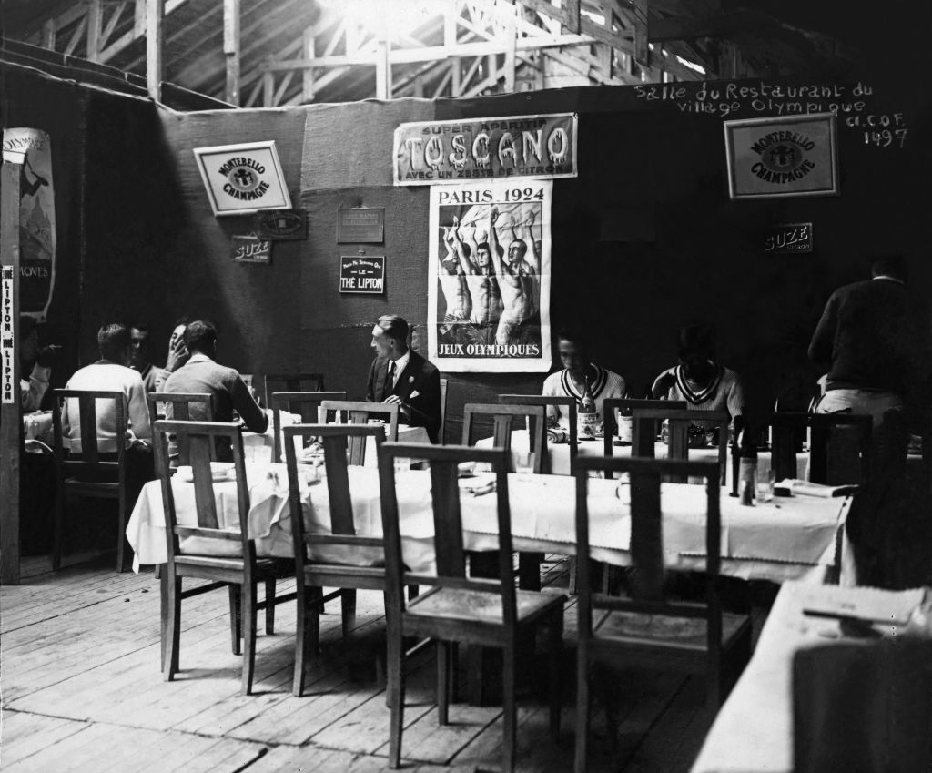 Several people are seated at tables in a vintage restaurant decorated with posters, one of which references the 1924 Paris Olympics