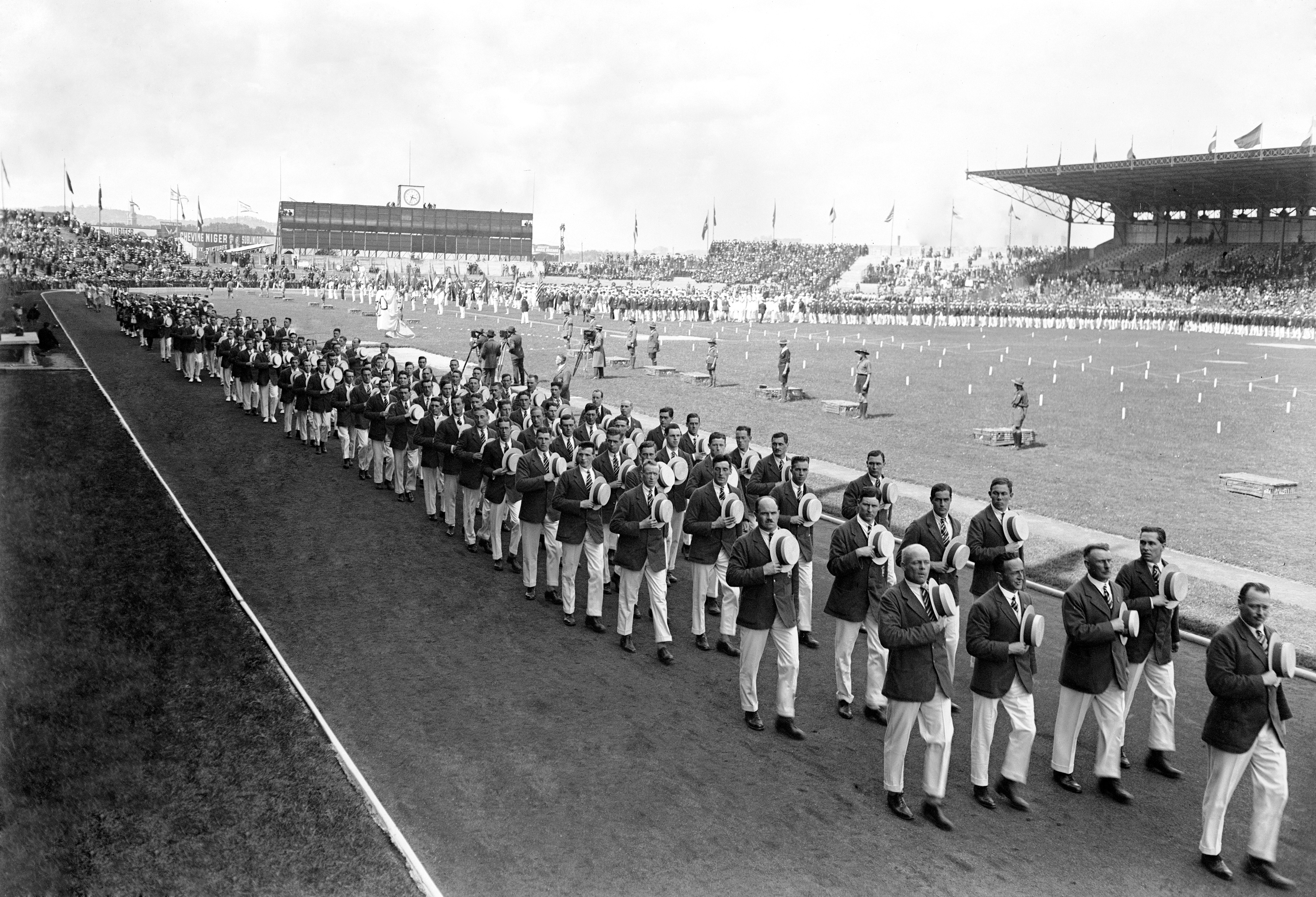 A large group of British athletes march in formation on a sports field during an opening ceremony in a stadium. Spectators fill the stands in the background