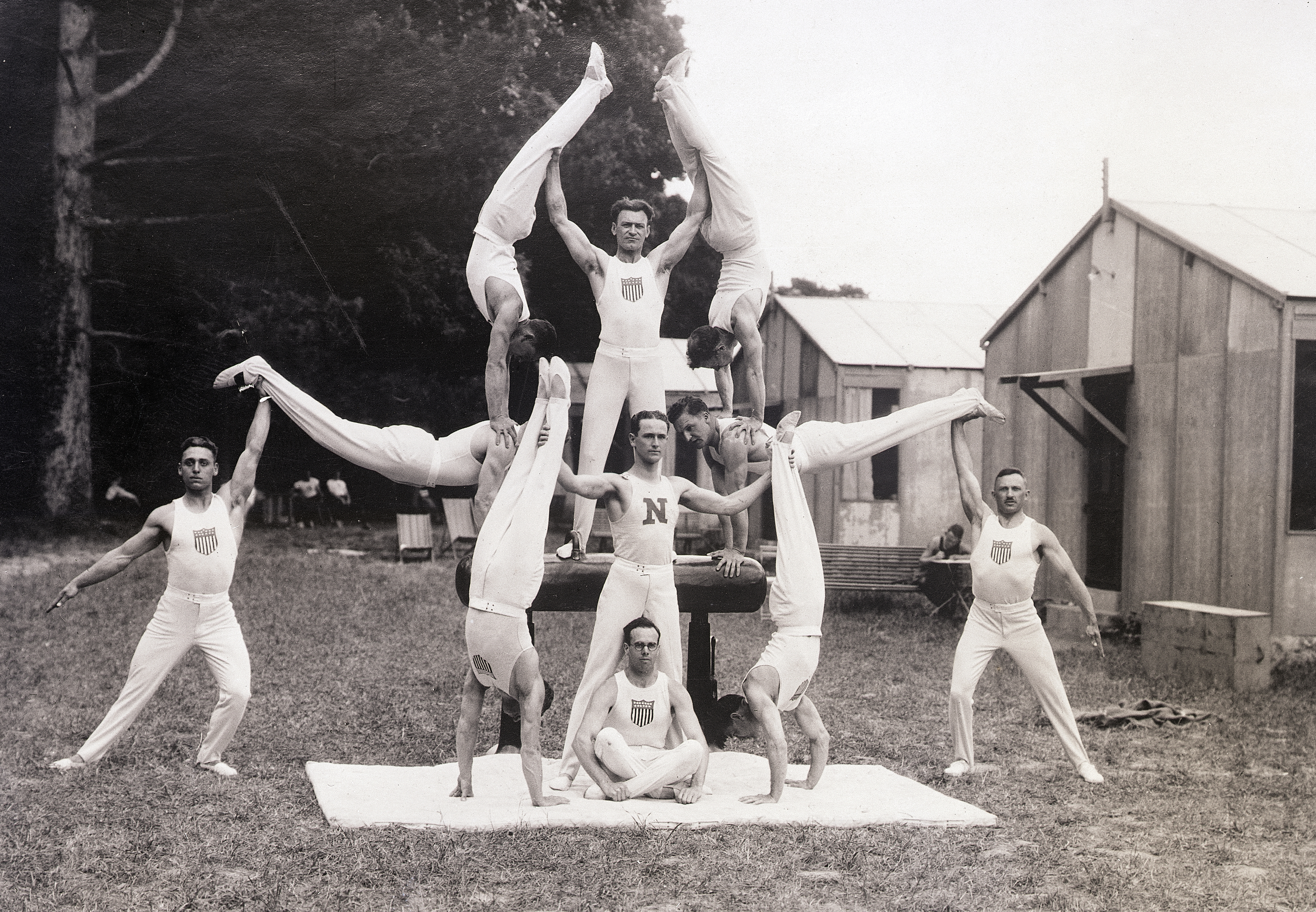 A group of nine male gymnasts perform an acrobatic pyramid outdoors, wearing matching sleeveless outfits with an emblem