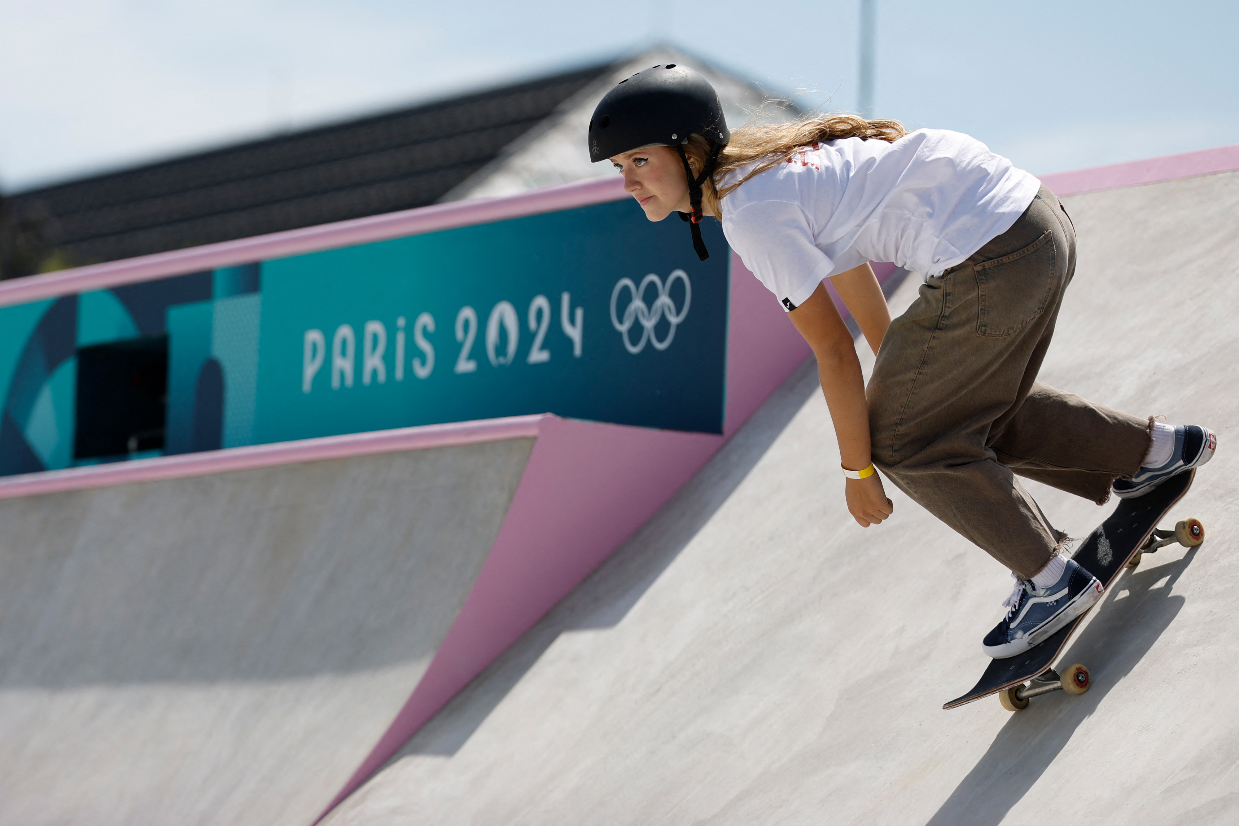 A young woman rides a skateboard on a ramp with &quot;Paris 2024&quot; and Olympic rings in the background. She is wearing a helmet, a white shirt, and brown pants