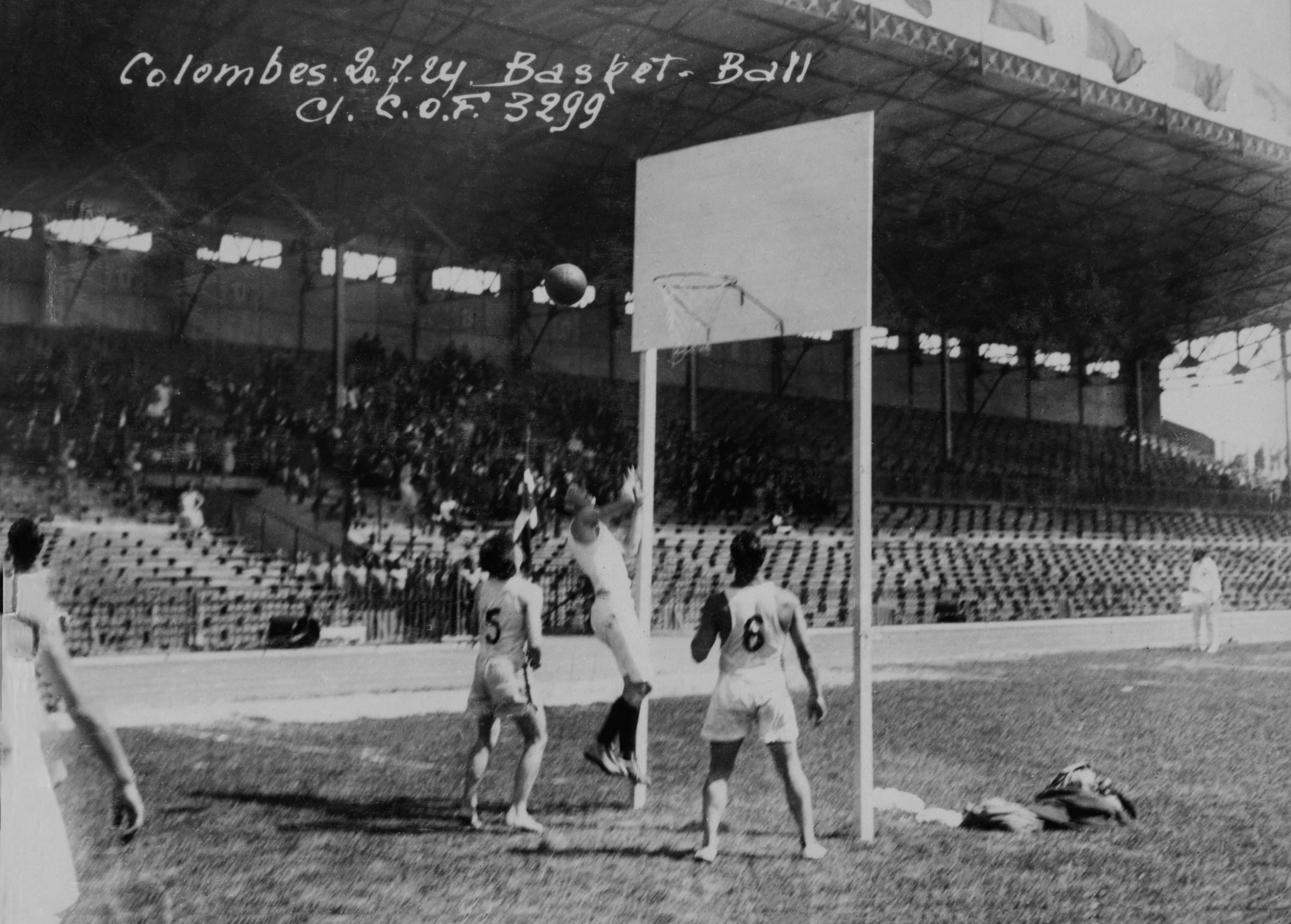 A historical outdoor basketball game is underway with four players in action under the hoop.