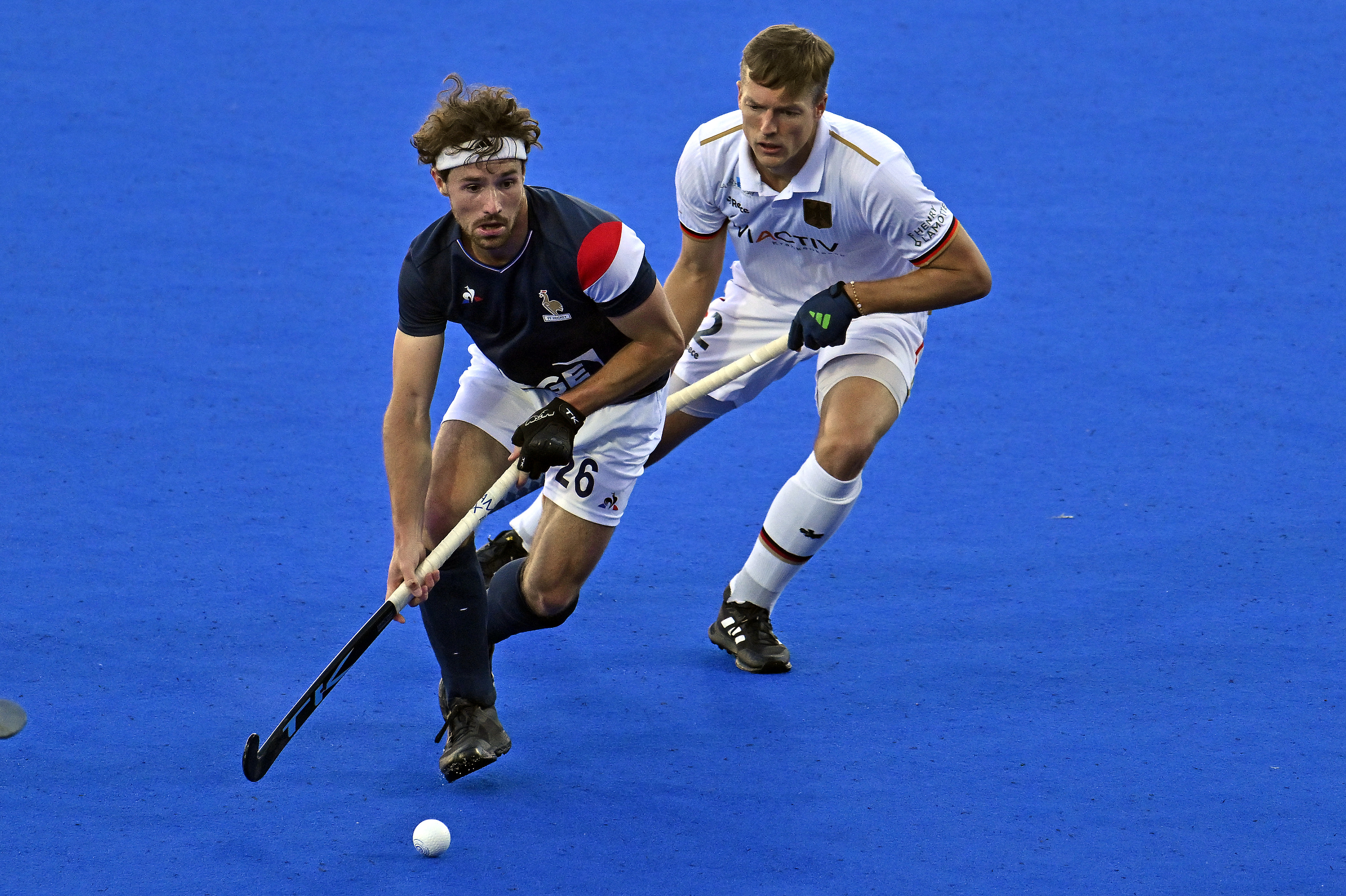 Two of France and Germany&#x27;s field hockey players in action on the blue pitch during a match, one about to hit the ball while another defends closely behind