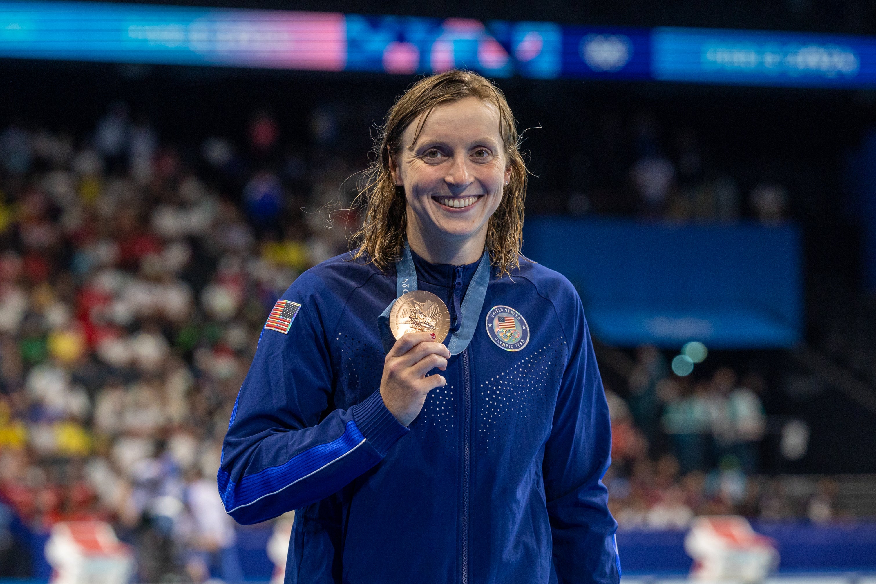 Katie Ledecky, wearing a sports jacket, holding up her bronze medal at a swimming competition