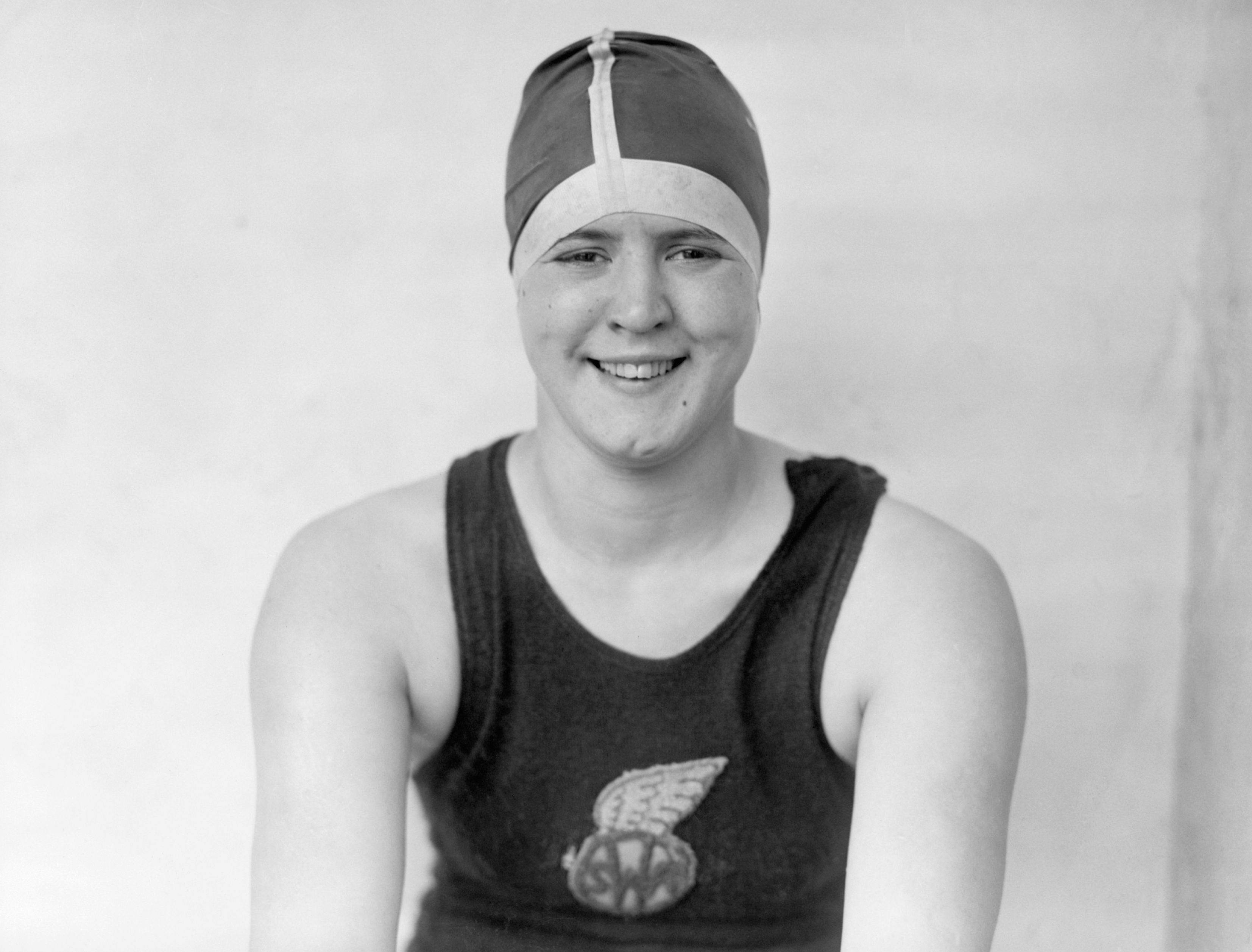 A swimmer smiles at the camera wearing a swim cap and a tank top with a winged emblem logo